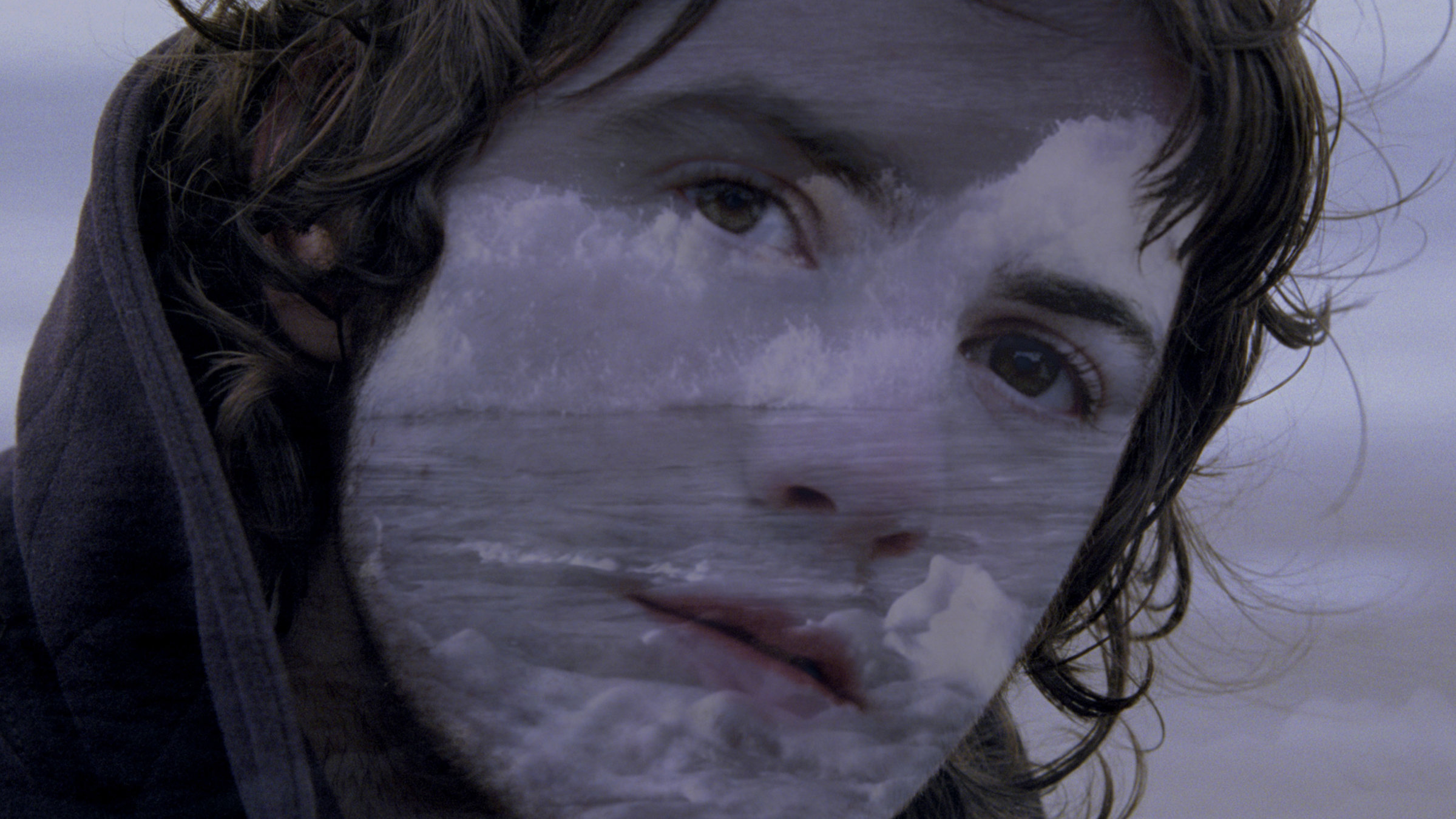 "Across the Universe": Kopf eines jungen Mannes mit dunklen Locken in Großaufnahme, in dessen Gesicht sich Gischt und Wellen spiegeln.