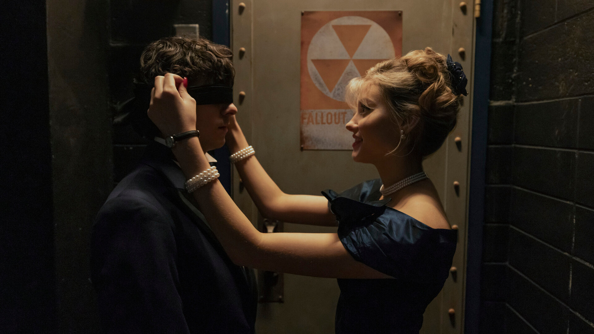 "Hysteria! - Schulball des Grauens": Judith (Jessica Treska) steht dicht vor Dylan (Emjay Anthony) und bindet ihm eine schwarze Augenbinde um. Die beiden befinden sich in einem düster beleuchteten Gang vor einer Metalltür mit einem Warnschild.