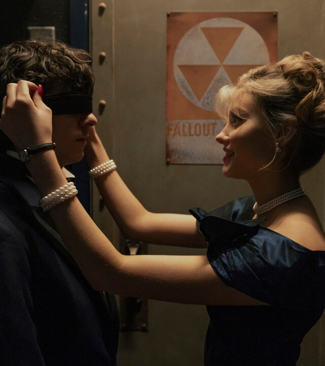 "Hysteria! - Schulball des Grauens": Judith (Jessica Treska) steht dicht vor Dylan (Emjay Anthony) und bindet ihm eine schwarze Augenbinde um. Die beiden befinden sich in einem düster beleuchteten Gang vor einer Metalltür mit einem Warnschild.