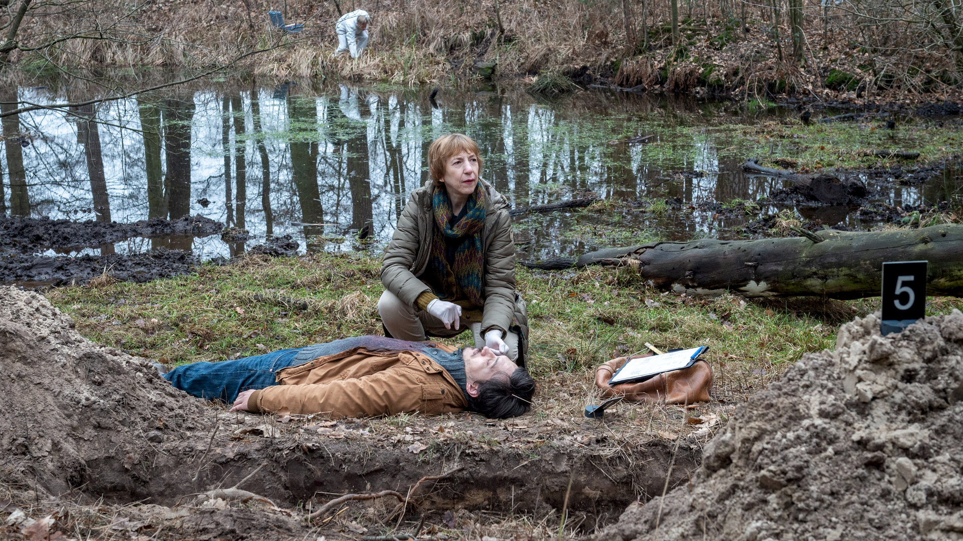 "SOKO Wismar – Welpenschutz": Daniel Kahlke (Mirko Krecar) liegt leblos auf dem Waldboden. Bei ihm kniet Helene (Katharina Blaschke) und führt ihre Untersuchungen durch. Ein großes Erdloch und Hügel von aufgeschütteter Erde deuten darauf hin, dass jemand den Mann vergraben wollte. Im Hintergrund liegen ein bemooster See und der Wald. Kriminaltechniker suchen nach Spuren am Ufer des Sees.