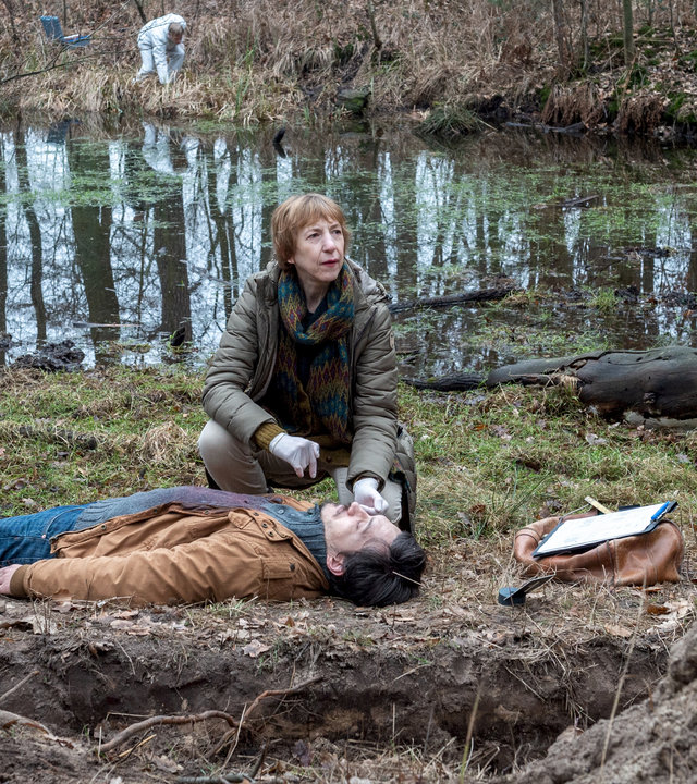 "SOKO Wismar – Welpenschutz": Daniel Kahlke (Mirko Krecar) liegt leblos auf dem Waldboden. Bei ihm kniet Helene (Katharina Blaschke) und führt ihre Untersuchungen durch. Ein großes Erdloch und Hügel von aufgeschütteter Erde deuten darauf hin, dass jemand den Mann vergraben wollte. Im Hintergrund liegen ein bemooster See und der Wald. Kriminaltechniker suchen nach Spuren am Ufer des Sees.