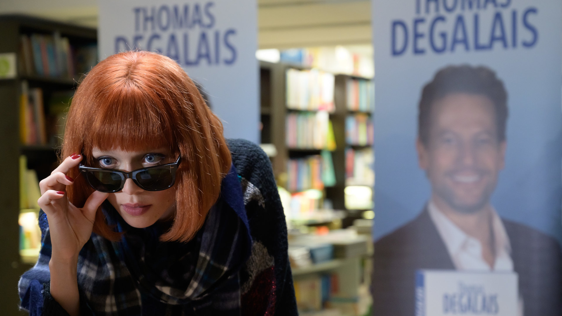 "Das Mädchen und die Nacht - Mord verjährt nicht": Pauline (Ivanna Sakhno) trägt eine rote Perücke. Sie schiebt ihre Sonnenbrille nach vorne, damit Thomas (Ioan Gruffudd) ihre Augen sehen kann. Im Hintergrund sind Plakate zu sehen, auf denen Thomas' Signierstunde angekündigt wird.