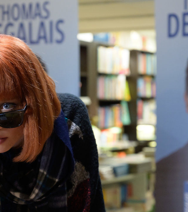 "Das Mädchen und die Nacht - Mord verjährt nicht": Pauline (Ivanna Sakhno) trägt eine rote Perücke. Sie schiebt ihre Sonnenbrille nach vorne, damit Thomas (Ioan Gruffudd) ihre Augen sehen kann. Im Hintergrund sind Plakate zu sehen, auf denen Thomas' Signierstunde angekündigt wird.
