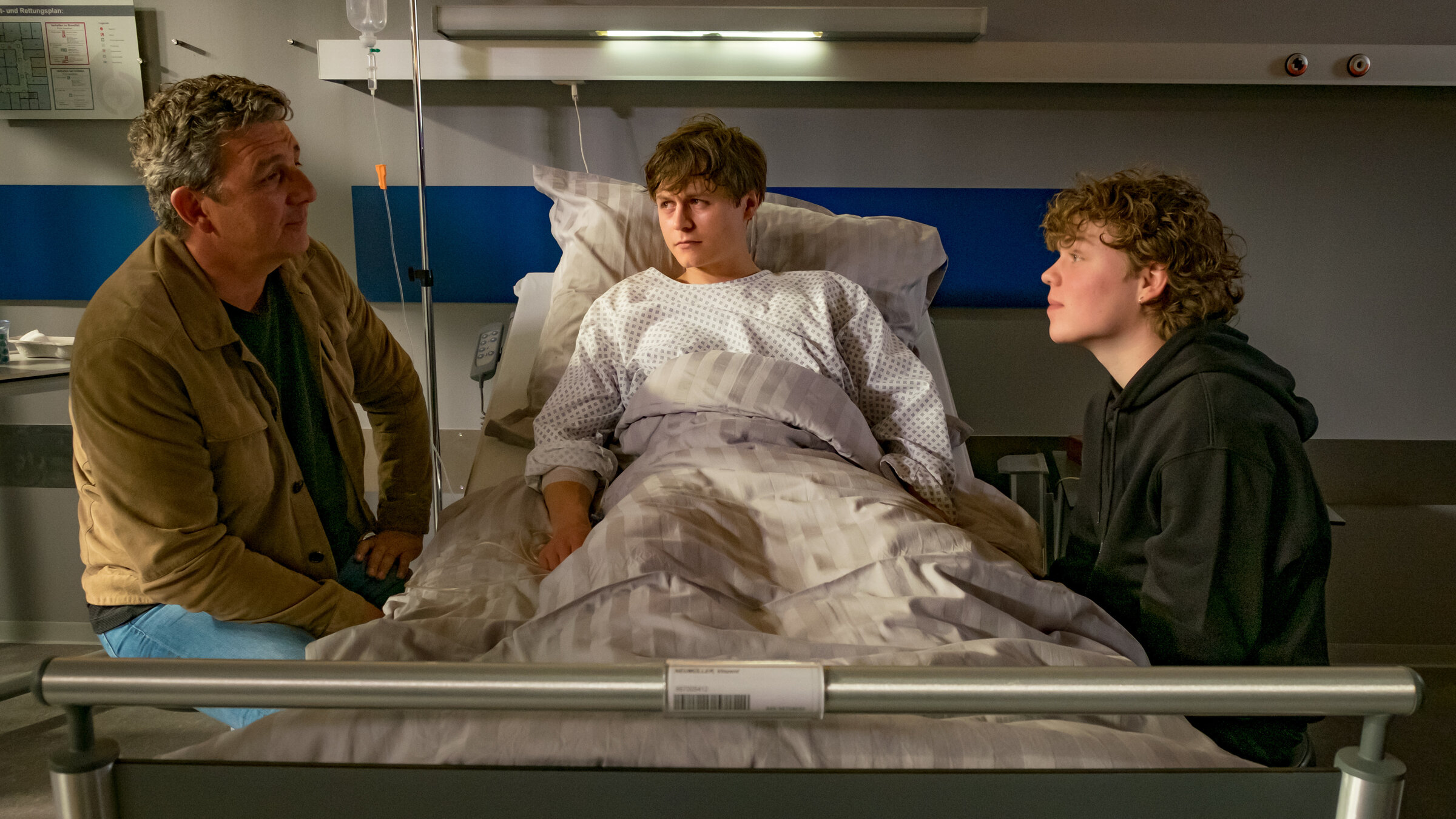 "Der Bergdoktor - Schuld": Im Krankenhaus: Mit blasser Gesichtsfarbe liegt Vinzent Neumüller (Tom Böttcher), ein Patientenhemd tragend, im Krankenbett. Rechts und links des Krankenbetts sitzen Dr. Martin Gruber (Hans Sigl) und Lennart Neumüller (Jonas Kaufmann). Sie blicken sich lächelnd an.