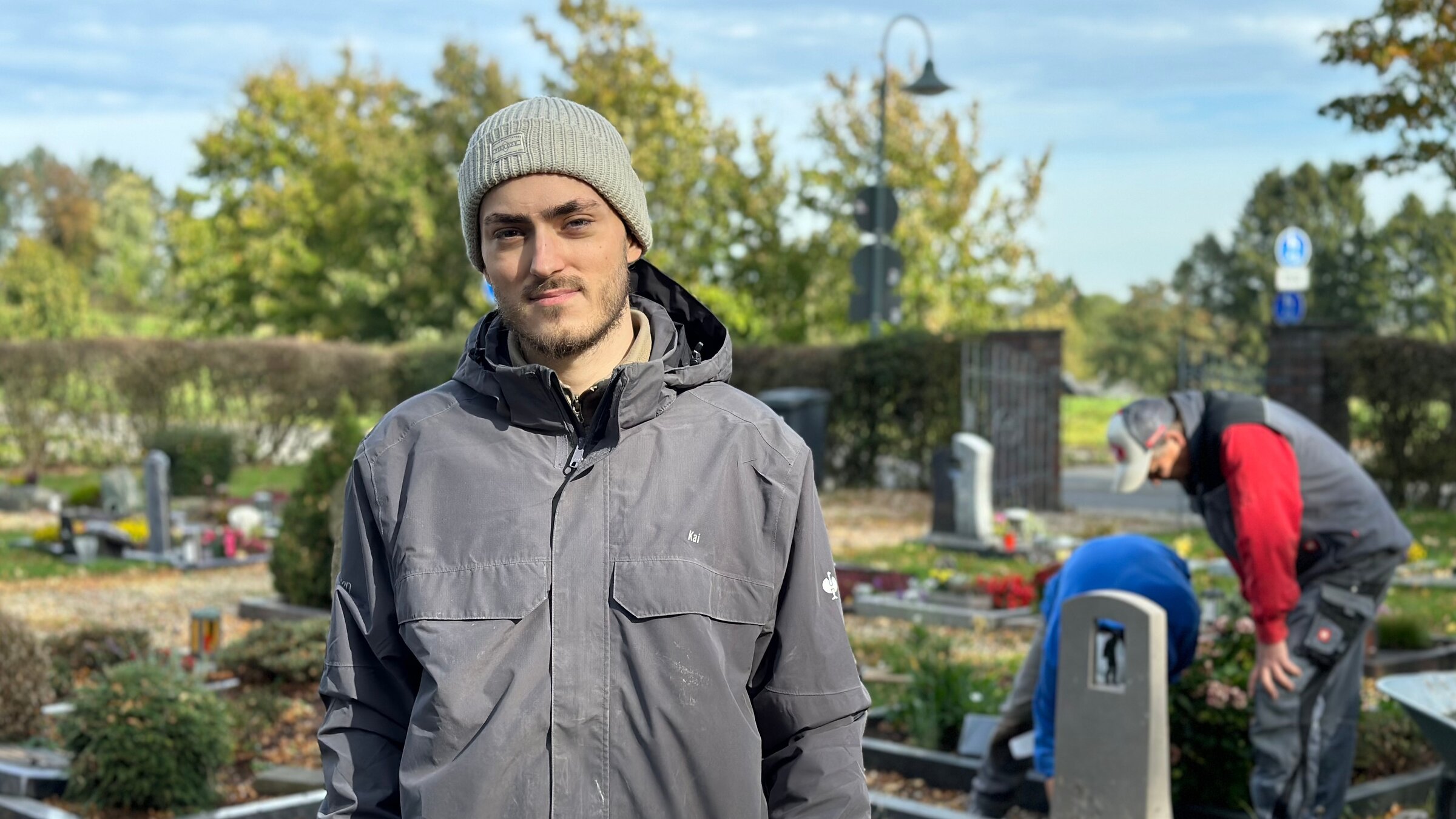 "ZDF.reportage: Bye-bye, Uni - Handwerk statt Hörsaal": Steinmetzlehrling Kai Niklas Kanehl steht vor Grabsteinen auf einem Friedhof. Im Hintergrund arbeiten sein Chef und ein weiterer Lehrling an einer Grabstelle.