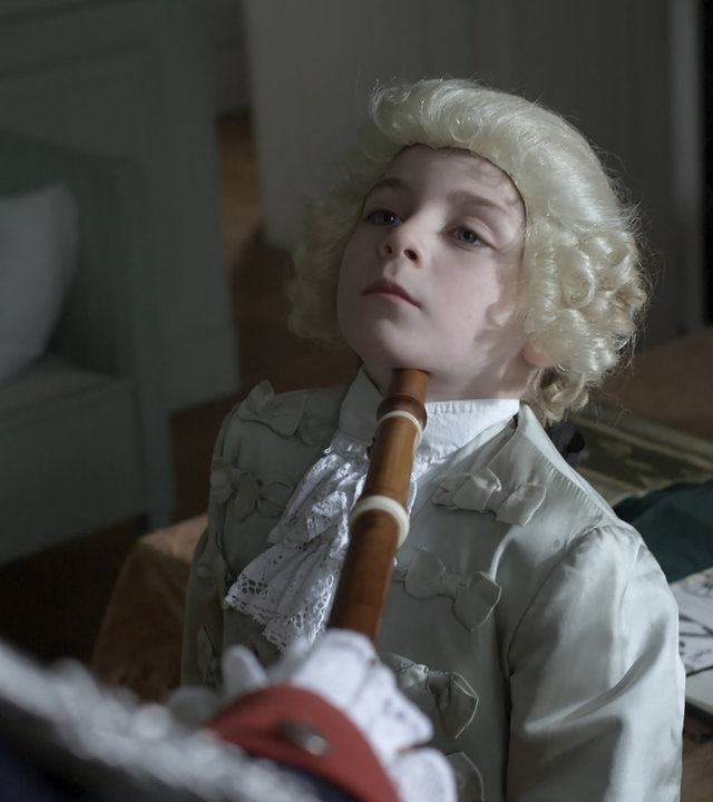 "Die glorreichen 10 - Der undankbarste Nachwuchs der Geschichte": Die Kamera schaut rechts an einem Mann mit Uniform vorbei auf einen kleinen Jungen in feinem Gewand und mit Lockenperücke. Der Mann hält dem Jungen eine Flöte unters Kinn, der deshalb sein Kinn weit anhebt.