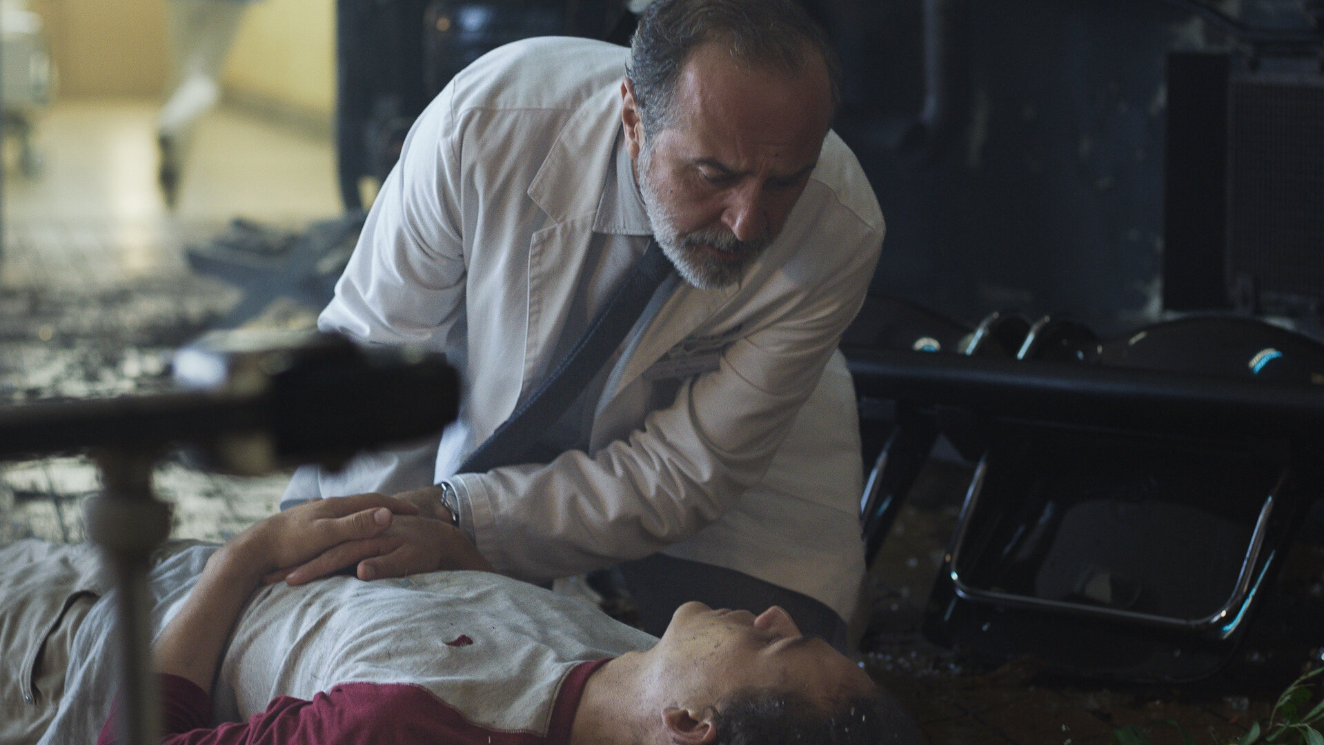 "Doktor Ballouz - Ein Sturm zieht auf": Juri (David Ali Rashed) liegt in einem Innenraum am Boden. Doktor Ballouz (Merab Ninidze) kniet neben ihm und hat eine Hand auf seinen Bauch gelegt. Beide schauen sich an.