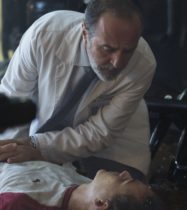 "Doktor Ballouz - Ein Sturm zieht auf": Juri (David Ali Rashed) liegt in einem Innenraum am Boden. Doktor Ballouz (Merab Ninidze) kniet neben ihm und hat eine Hand auf seinen Bauch gelegt. Beide schauen sich an.