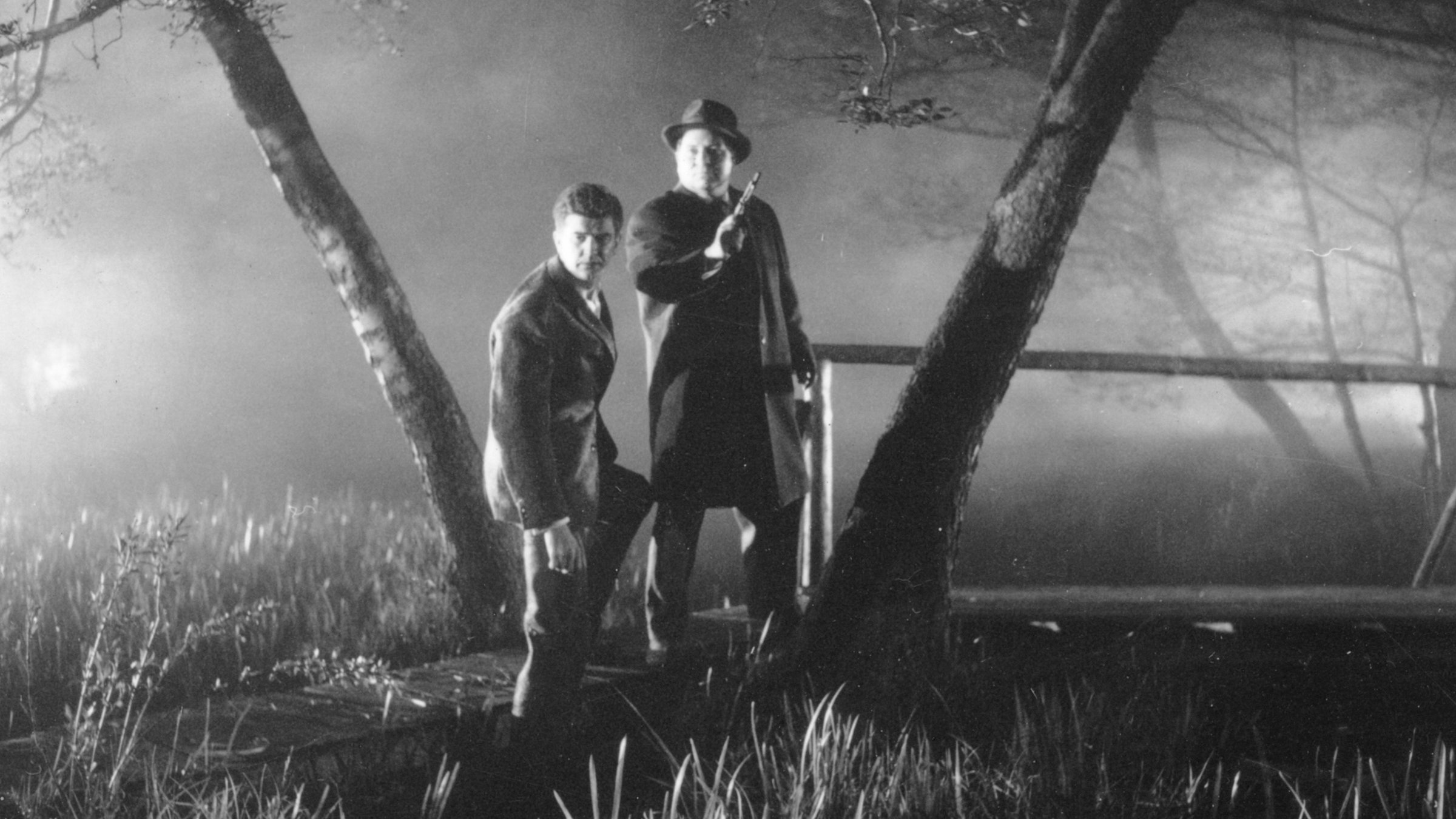 “Der Würger von Schloss Blackmoor“: Inspektor Jeff Mitchell (Harry Riebauer) und sein Assistent Watson stehen am Ende einer Brücke zwischen zwei Bäumen und blicken zielgerichtet geradeaus. Dabei hält Watson eine nach oben gerichtete Schusswaffe in seiner Hand.