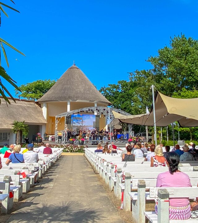"Evangelischer Open-Air Gottesdienst - Gastfreundschaft": Der Konzertgarten des Ostseebads Kühlungsborn liegt unter einem strahlend blauem Himmel, umsäumt von Palmen. Die Zuhörenden sitzen auf weißen Holzbänken und blicken, von der Kamera abgewandt, nach vorne zur Bühne.