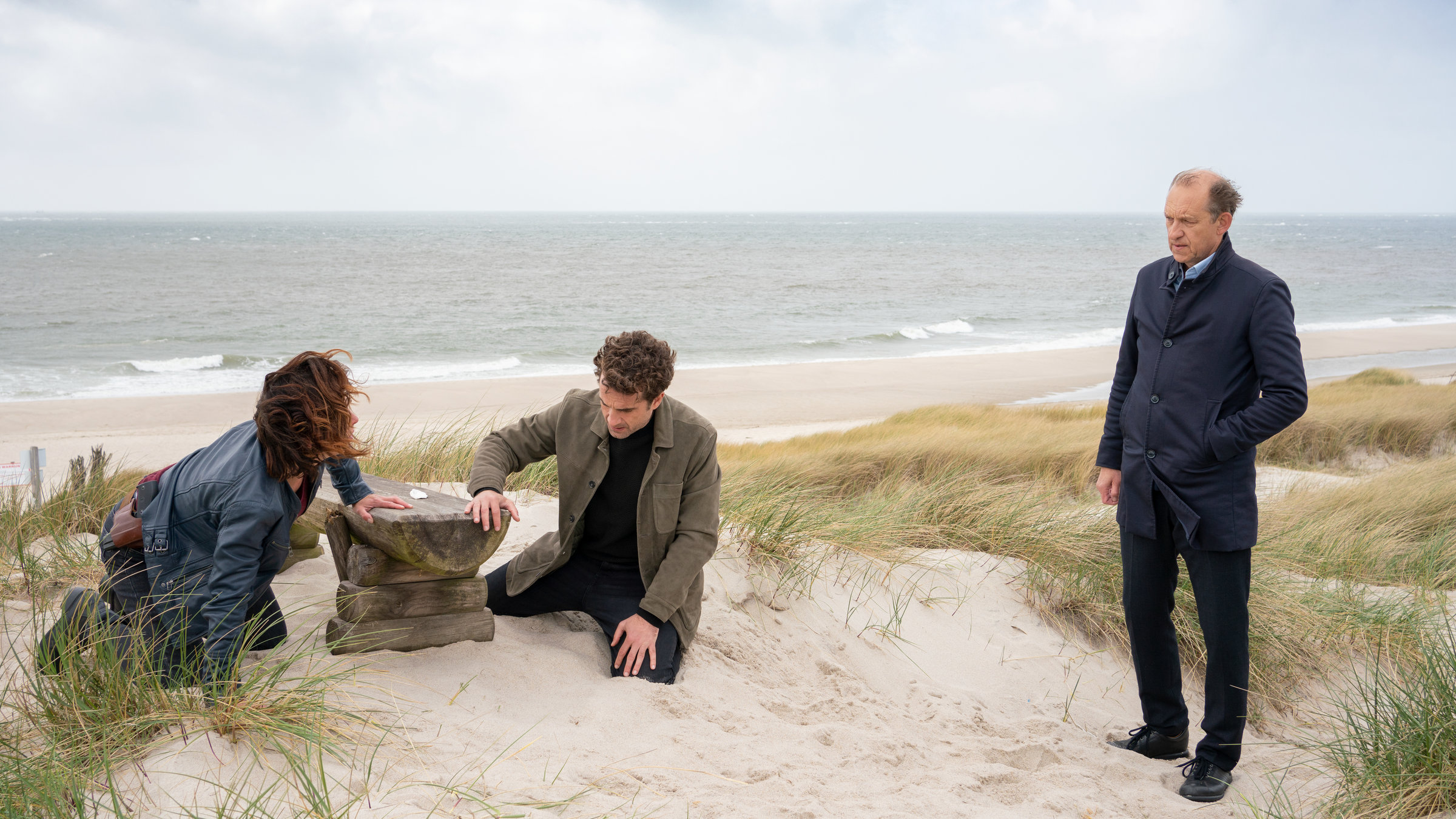 "Nord Nord Mord - Sievers und der erste Schrei": Ina Behrendsen (Julia Brendler), Hinnerk Feldmann (Oliver Wnuk) und Carl Sievers (Peter Heinrich Brix) sind auf einer Düne direkt am Strand von Sylt. Sie suchen etwas im Sand unter einer Bank zwischen dem Seegras. Ina und Feldmann knien an der Holzbank, Sievers schaut ihnen mit einer Hand in der Manteltasche zu. Im Hintergrund ist der breite Strand mit dem offenen Meer zu sehen.