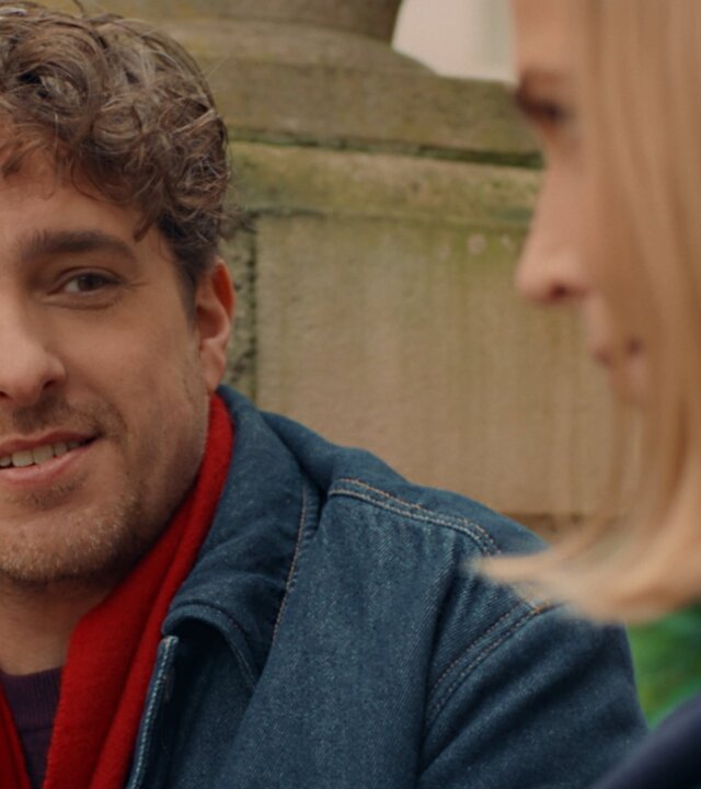 "Single Bells - 12 Dates till Christmas: Fonny": Fonny (Jelle De Beule) wirft Mina (Julie Van den Steen) einen flirtenden Blick zu. Er hat gelocktes Haar und trägt einen Dreitagebart.