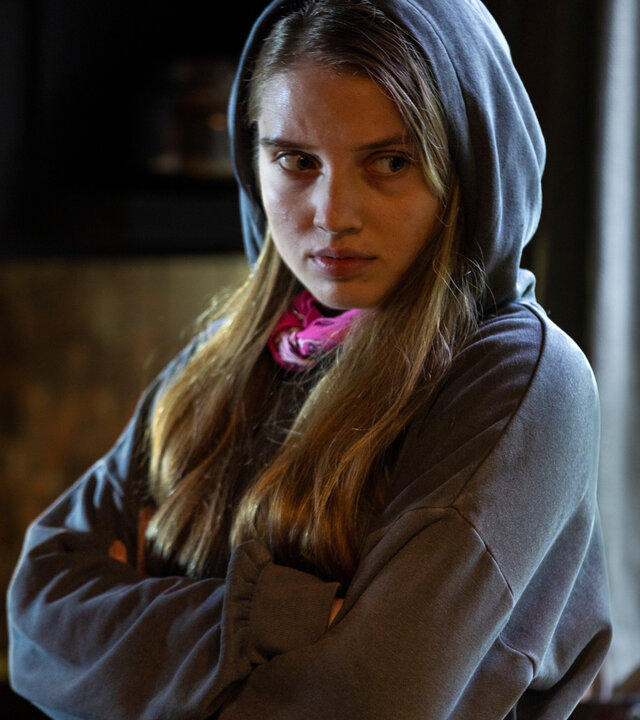 "The Kollective - Quid pro quo": Anna (Veronika Mox) steht mit verschränkten Armen in einem Raum mit dem Rücken zu einem Fenster. Sie trägt einen Hoodie und wirkt verärgert.