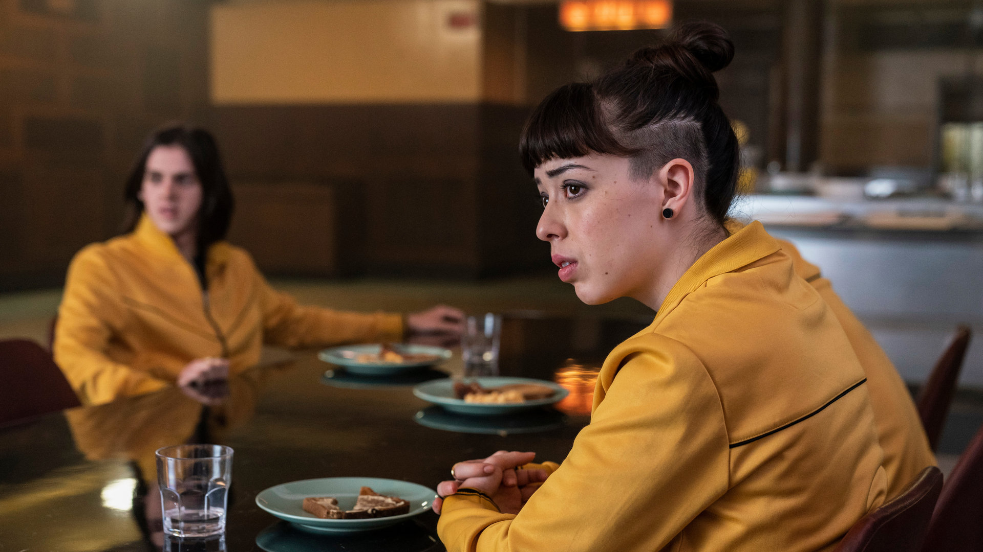 "Alex Rider - Fluchtplan": Kyra (Marli Siu) sitzt in der Kantine an einem Tisch und blickt traurig zur Seite. Gegenüber von ihr sitzt James (Earl Cave) und schaut in dieselbe Richtung. Sie tragen beide einen gelben Pullover.