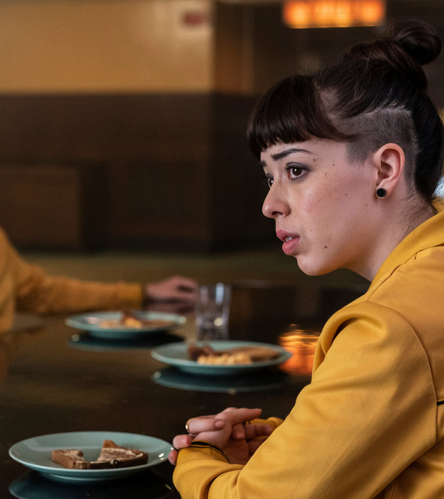 "Alex Rider - Fluchtplan": Kyra (Marli Siu) sitzt in der Kantine an einem Tisch und blickt traurig zur Seite. Gegenüber von ihr sitzt James (Earl Cave) und schaut in dieselbe Richtung. Sie tragen beide einen gelben Pullover.