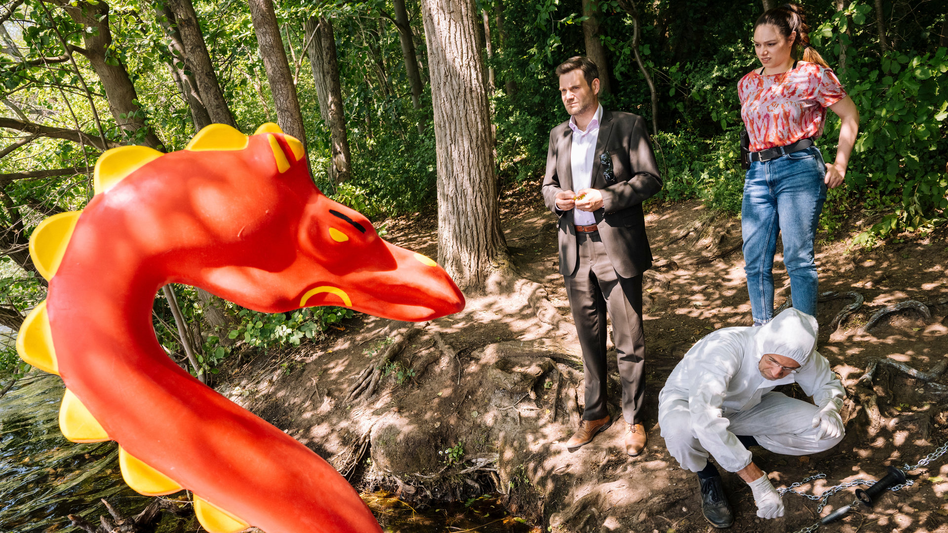 "SOKO Köln - Der Sturm op et Rodhuus": Jonas Fischer (Lukas Piloty) steht vor Amira Mahdi (Aylin Ravanyar) in einem Wald am Ufer eines Sees und blickt nachdenklich auf einen orangenen, aufgeblasenen Drachen, der vor ihnen steht. Amira sieht den Drachen ebenfalls verwirrt an. Vor ihr kniet ein Mitarbeiter der Spurensicherung (Komparse) auf dem Waldboden.