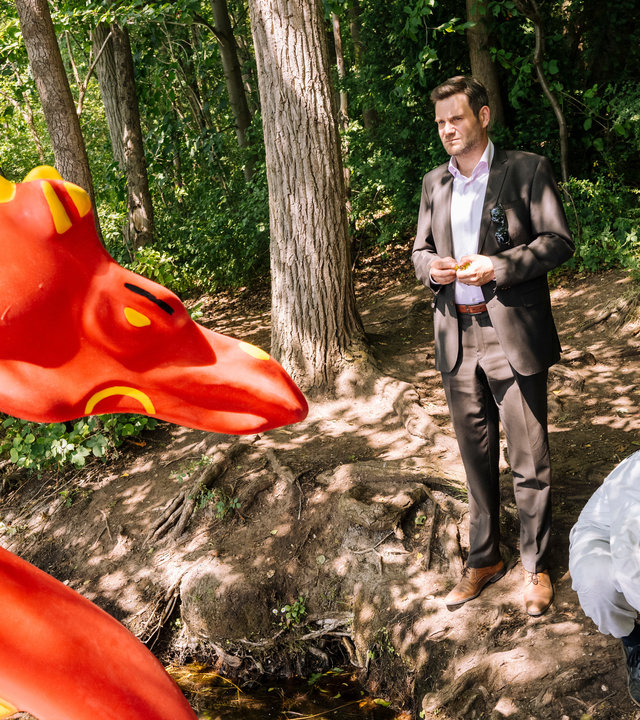 "SOKO Köln - Der Sturm op et Rodhuus": Jonas Fischer (Lukas Piloty) steht vor Amira Mahdi (Aylin Ravanyar) in einem Wald am Ufer eines Sees und blickt nachdenklich auf einen orangenen, aufgeblasenen Drachen, der vor ihnen steht. Amira sieht den Drachen ebenfalls verwirrt an. Vor ihr kniet ein Mitarbeiter der Spurensicherung (Komparse) auf dem Waldboden.