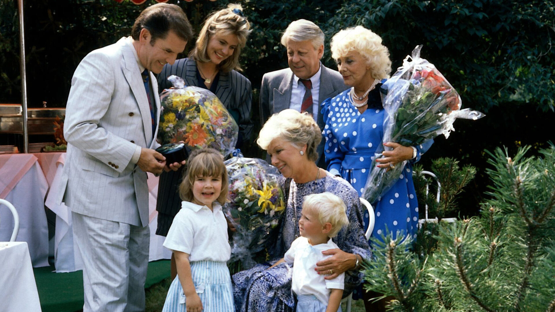 "Die Wicherts von nebenan: Hannelore wird 50": In einem Garten. Hannelore (Maria Sebaldt) hat Geburtstag und steht im Kreise ihrer Familie. Sie schaut liebevoll auf ihre beiden Enkelkinder. Drei der Gratulanten haben Blumensträuße in der Hand.