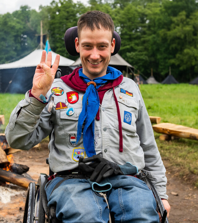 "einfach Mensch - Lion Broschat: Ich bin ein Pfadfinder": Lion Broschat sitzt im Rollstuhl vor einem brennenden Feuer an einer Feuerstelle. Er trägt eine graue Pfadfinderkluft und ein blaues Halstuch und lacht in die Kamera. Im Hintergrund steht ein großes schwarzes Zelt neben einem Zelt mit einem weißen Dach auf einer Wiese vor Bäumen.