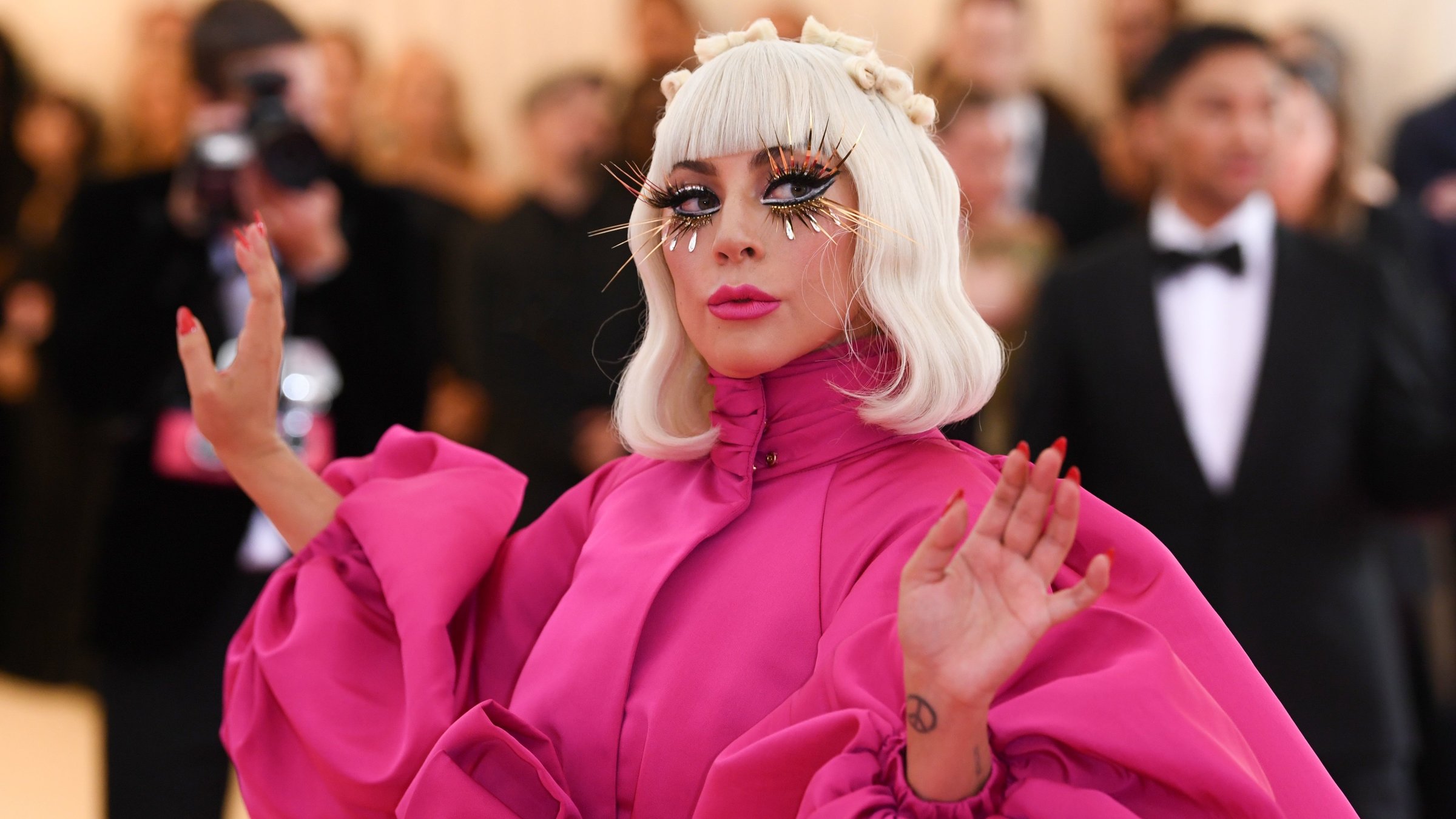 "The True Story of Lady Gaga": Lady Gaga trägt ein rosafarbenes Kleid mit Puffärmeln. Hellblonde Haare reichen ihr bis zur Schulter, und sie trägt lange künstliche Wimpern. Im Hintergrund sind Fotografen und ein Mann im Smoking zu sehen.