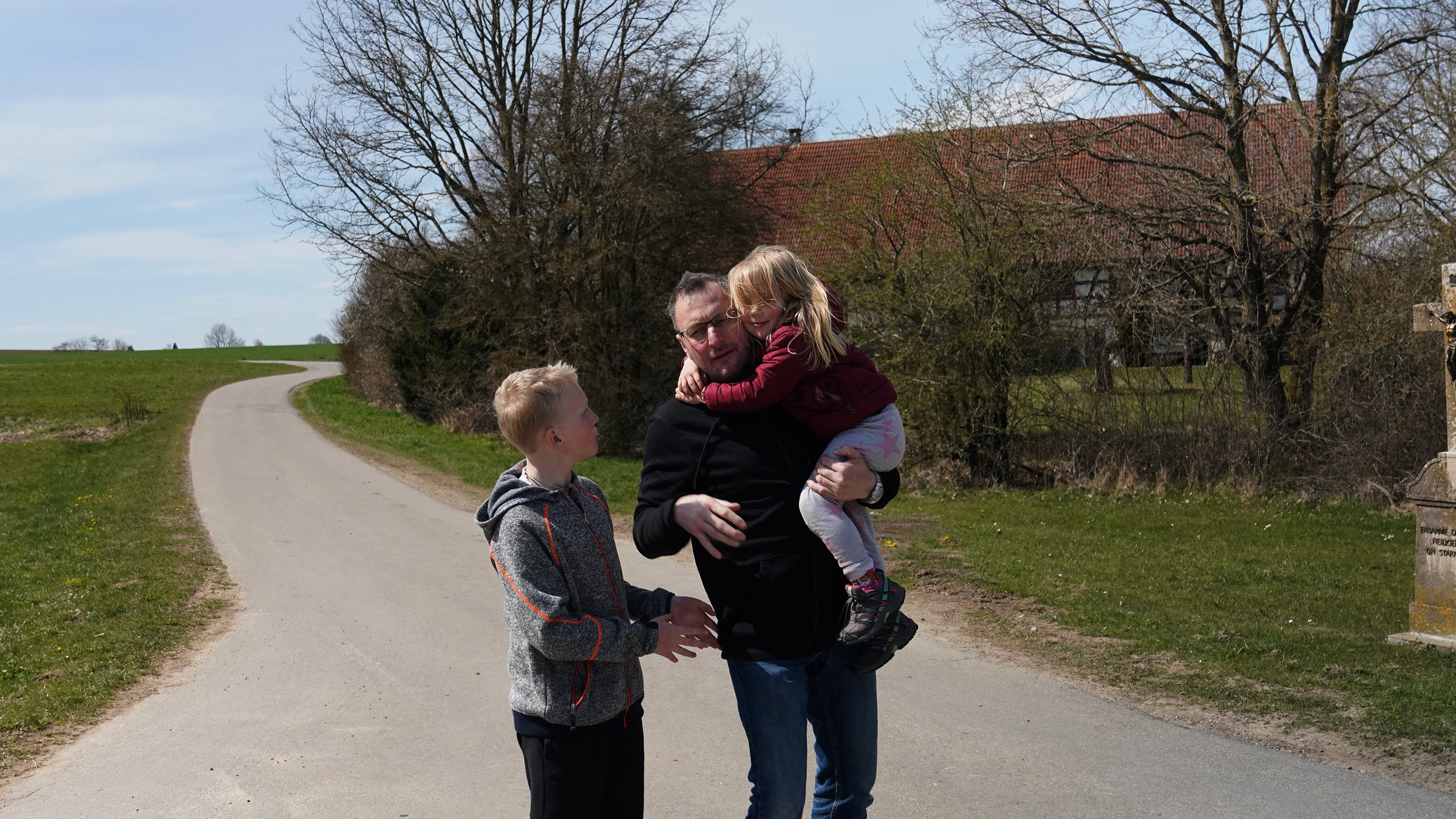 "37° Jung, verwitwet und allein": Stephan steht mit sein beiden Kindern auf einem gepflasterten Weg, links ist ein großes Feld und rechts ein Gebäude. Stephan trägt das Mädchen auf dem Arm. Der größere Junge steht neben ihm.
