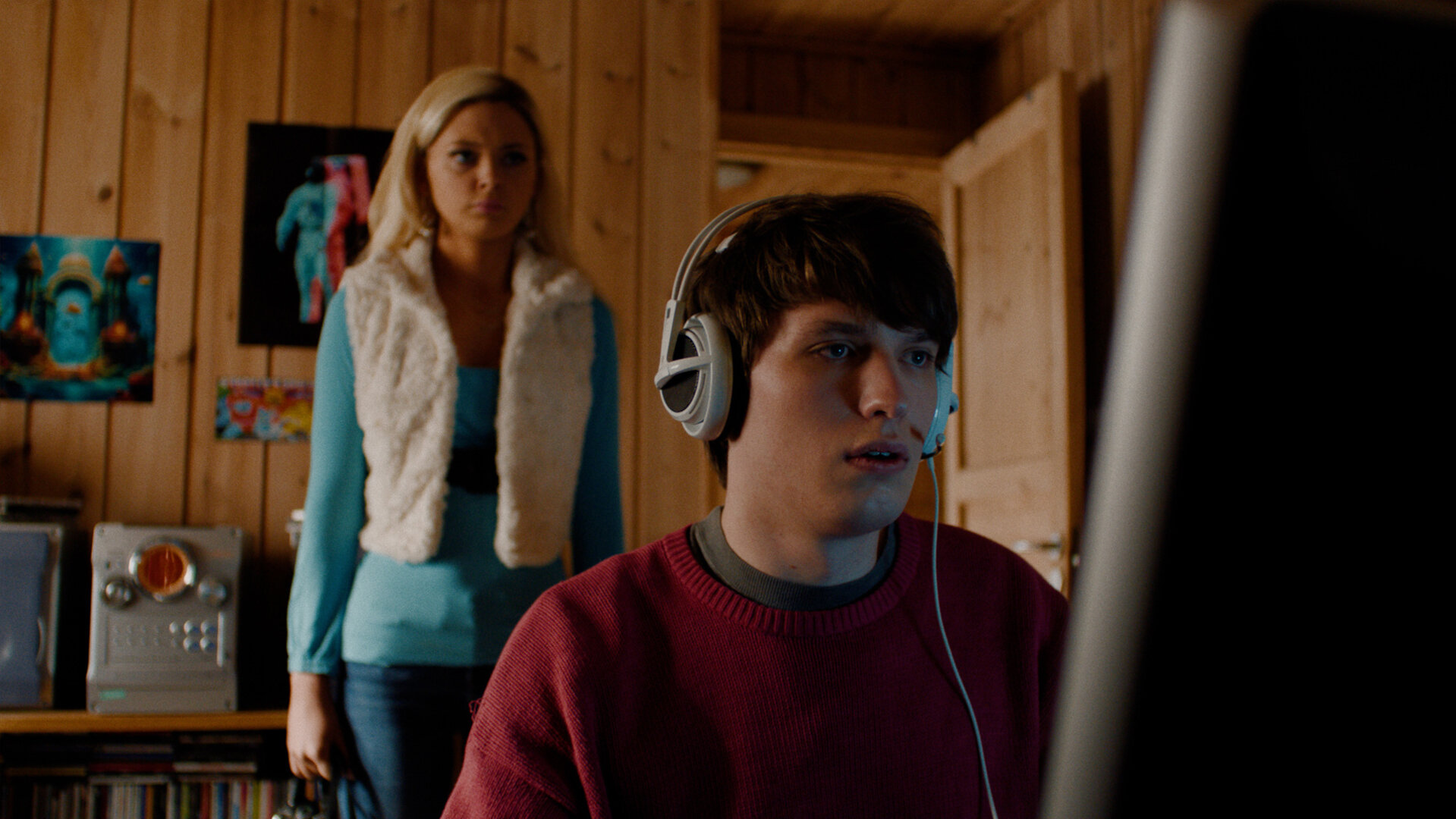 "Requiem for Selina - Schmetterlings-Effekt": Johan (Oliver Sletvold Andersen) sitzt mit einem Headset auf den Ohren in seinem Zimmer und schaut auf seinen Computer-Bildschirm. Hinter ihm steht Selina (Elli Rhiannon Müller Osborne) und schaut ihn an.