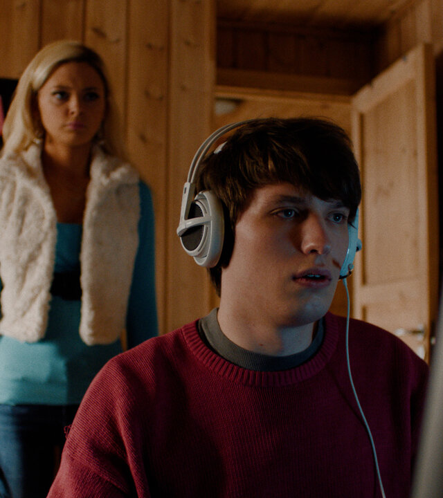 "Requiem for Selina - Schmetterlings-Effekt": Johan (Oliver Sletvold Andersen) sitzt mit einem Headset auf den Ohren in seinem Zimmer und schaut auf seinen Computer-Bildschirm. Hinter ihm steht Selina (Elli Rhiannon Müller Osborne) und schaut ihn an.