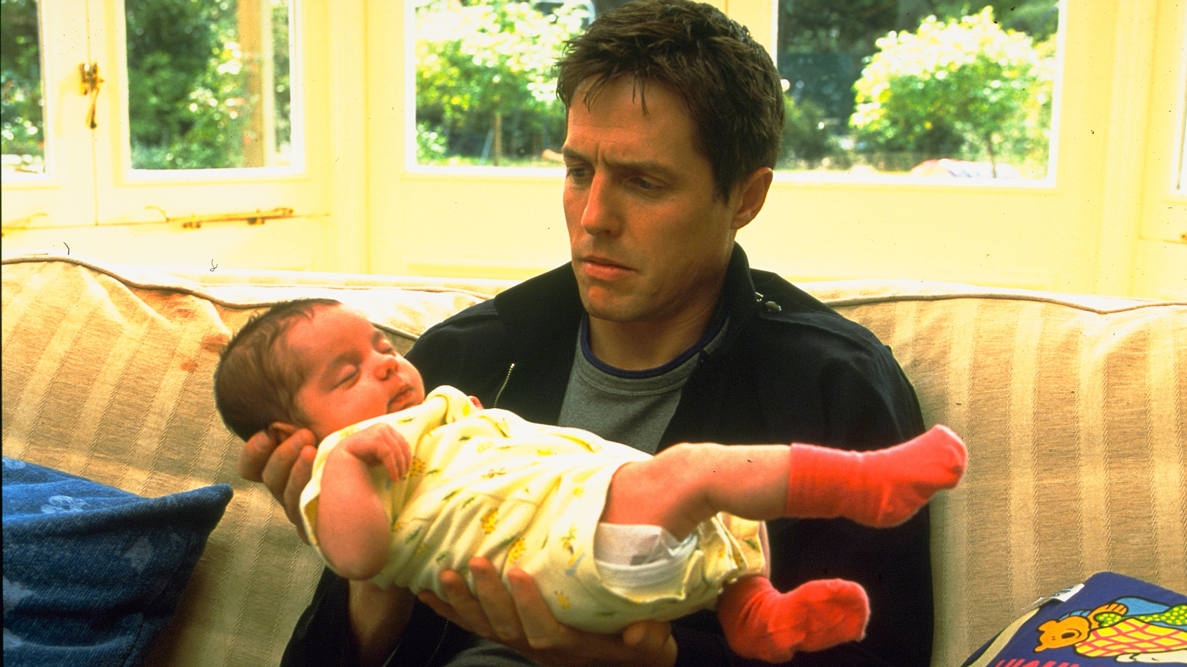 "About a Boy oder: Der Tag der toten Ente": Will (Hugh Grant) sitzt auf einem Sofa in einem sonnigen Raum und hält ein Baby mit beiden Händen vor sich. Äußerst skeptisch betrachtet er das Baby.