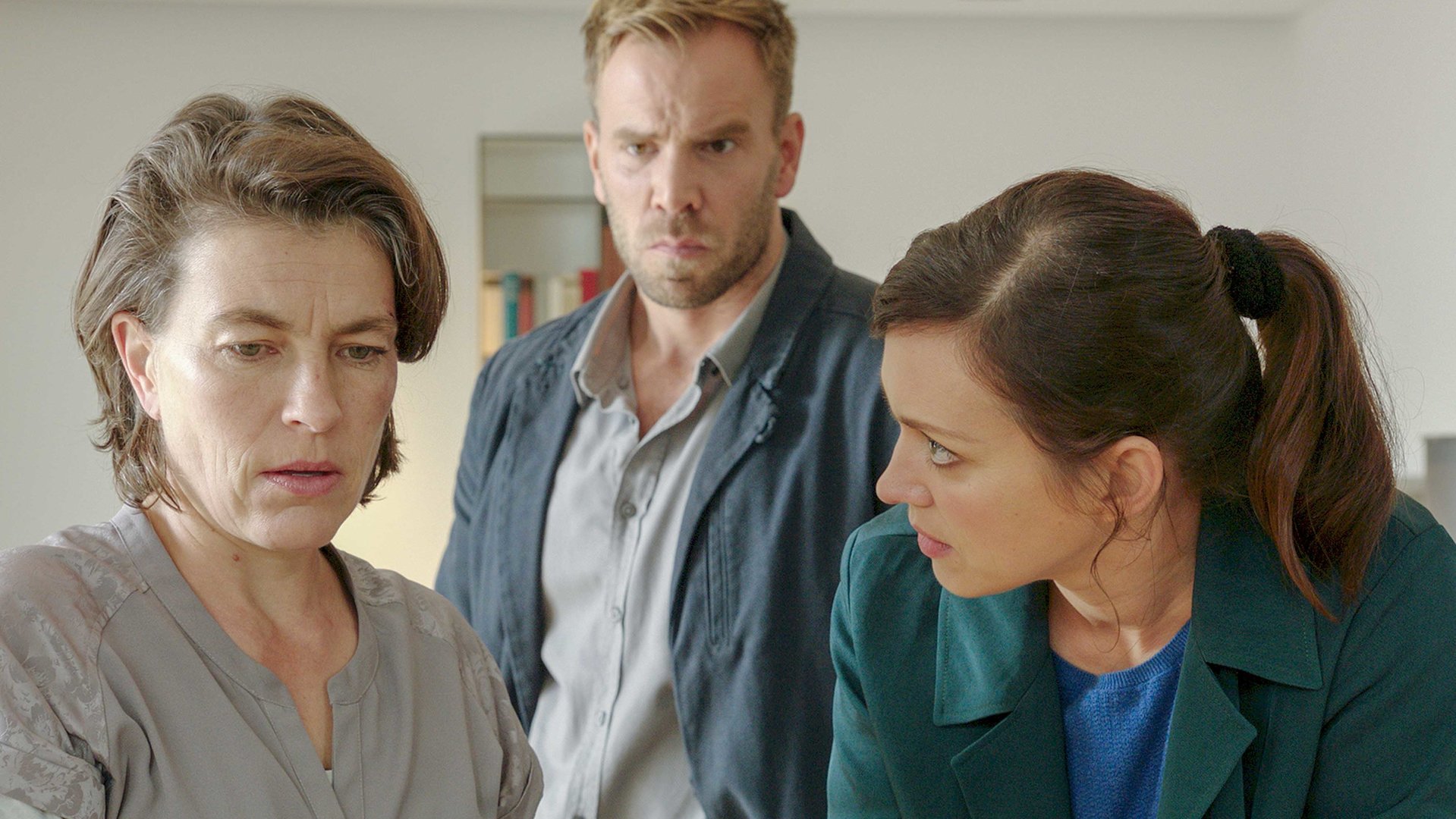 "SOKO Wismar – Aus Liebe": Juliane Schöller (Jule Gartzke) wird von der Kommissarin Karoline Joost (Nike Fuhrmann) befragt. Sie erwidert den Blick der Kommissarin nicht, sondern guckt ins Leere. Hinter den beiden steht Kommissar Lars Pöhlmann (Dominic Boeer), der ernst zu Juliane Schöller sieht.