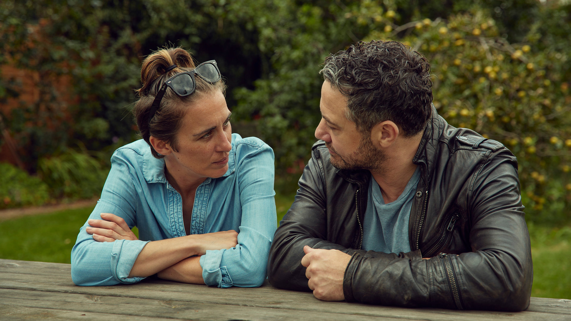 "Trigger Point: Hilferuf": Lana (Vicky McClure) und Karl (Warren Brown) sitzen draußen im Grünen nebeneinander an einem Holztisch, stützen sich mit den Armen auf und schauen sich an.