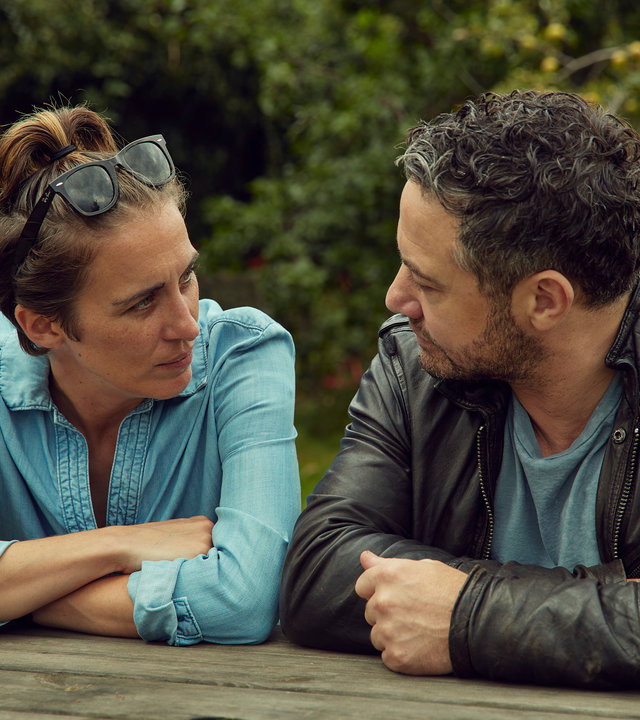 "Trigger Point: Hilferuf": Lana (Vicky McClure) und Karl (Warren Brown) sitzen draußen im Grünen nebeneinander an einem Holztisch, stützen sich mit den Armen auf und schauen sich an.