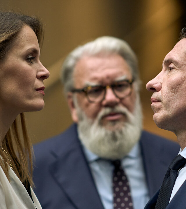 "Elixir – The Pain Killers - Aspergon": Isabelle (Hanna Verboom) und Juliën (Jacob Derwig) stehen sich gegenüber und schauen sich mit ernster Miene an. Etwas im Hintergrund steht Ludo (Wim Opbrouck) und beobachtet die beiden.