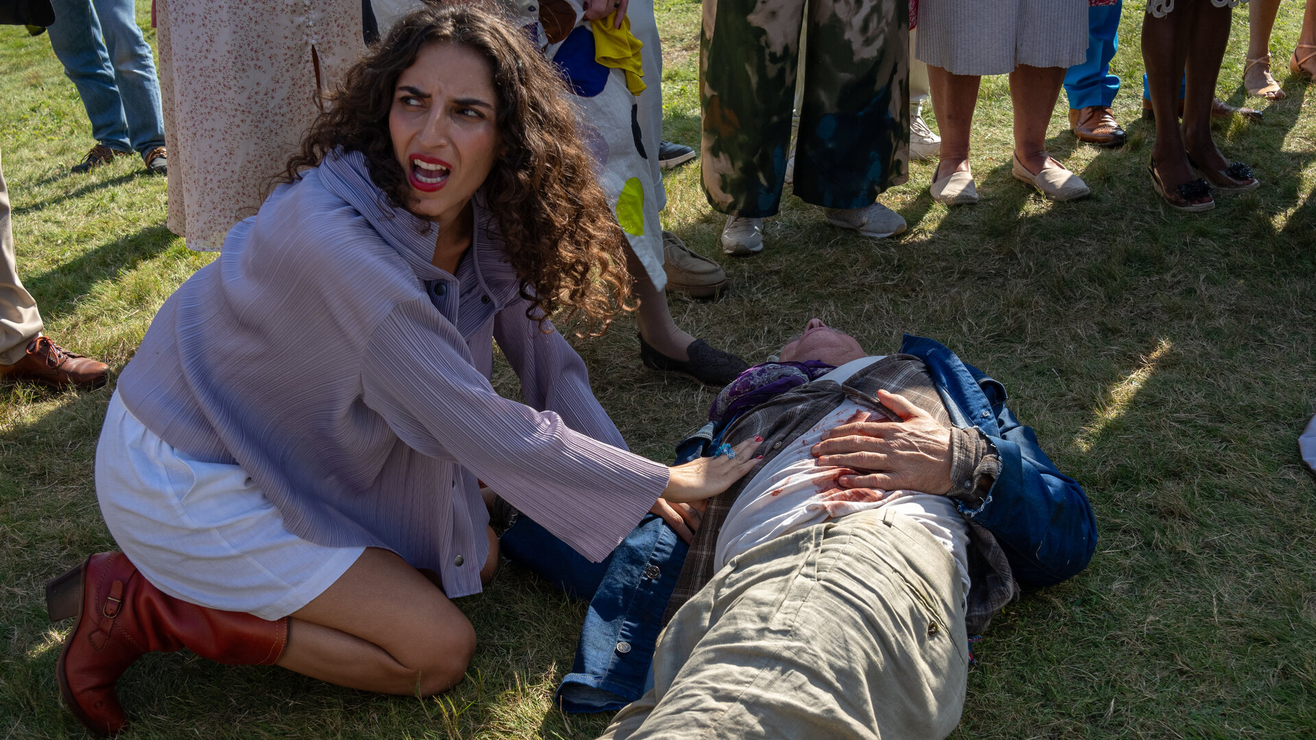 "Dan Sommerdahl - Tödliche Idylle: Der Patriarch": Daniella Rossing (Ellaha Lack) kniet neben dem im Gras liegenden Thor (Komparse), der sich die Hand auf eine blutende Wunde am Bauch hält. Daniella hat ihre Hände sanft auf ihn gelegt, blickt über ihre rechte Schulter und ruft verzweifelt um Hilfe.