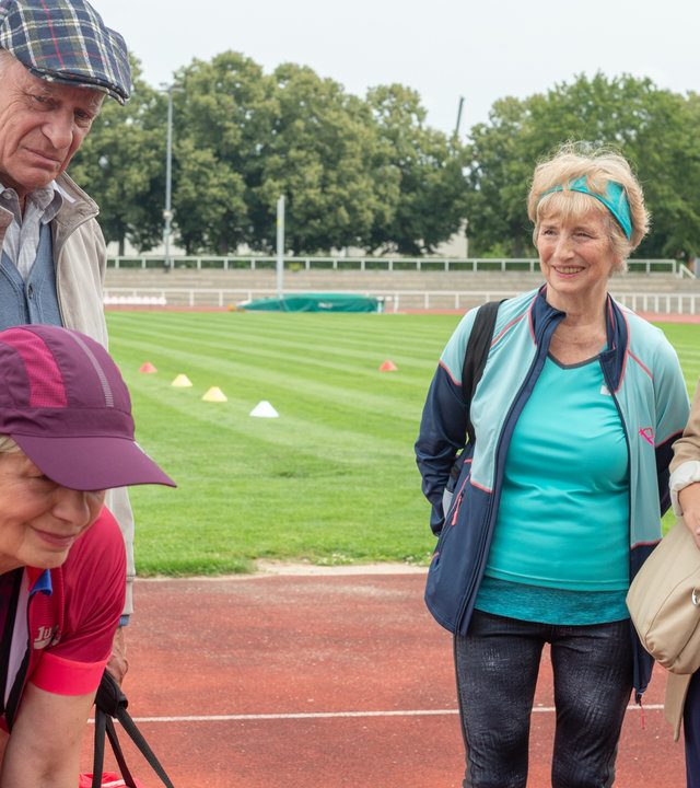 „SOKO Wismar – Tödliche Höhe“: Elisabeth Mayer (Christine Heinze) bindet ihre Schuhe zusammen und lächelt ein triumphierendes Lächeln. Hinter ihr stehen Dieter Mayer (Rüdiger Kuhlbrodt), der griesgrämig dreinschaut und Marianne (Dorothea Hagena) sowie Ellen Stegemann (Lisa Adler). Im Hintergrund trainieren Senioren auf dem Sportplatz.