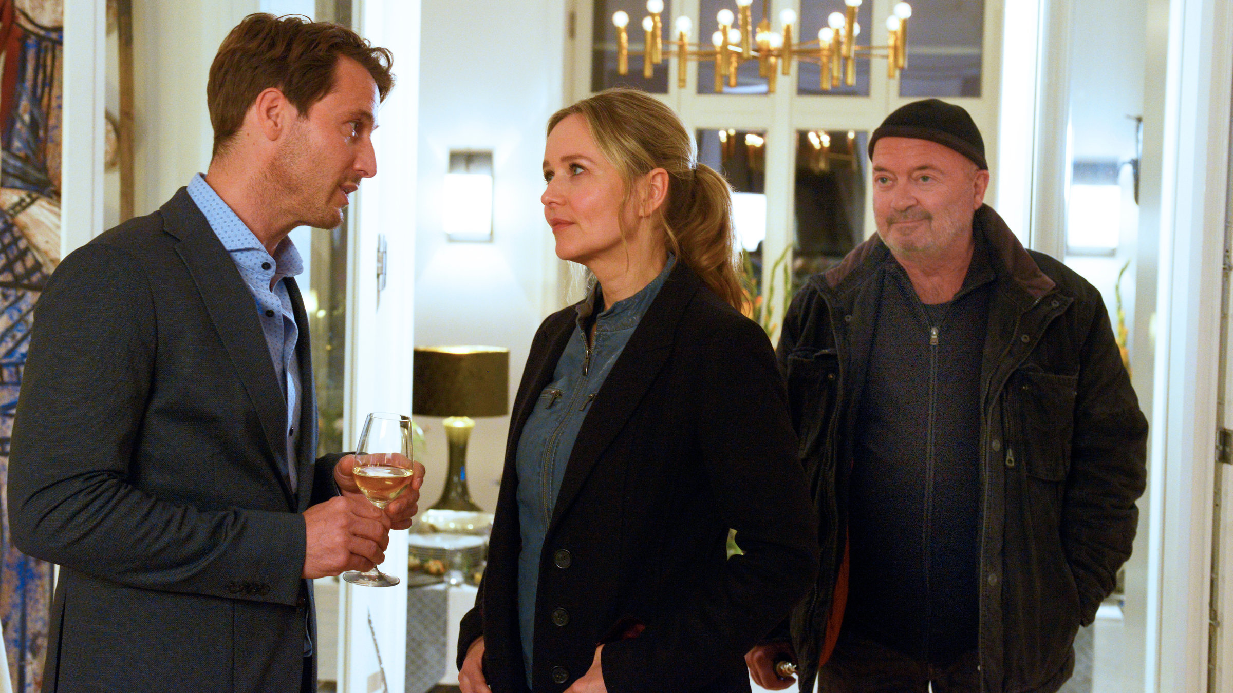 "Ein starkes Team - Schulzeit": Linett Wachow (Stefanie Stappenbeck) und Otto Garber (Florian Martens)stehen Hendrik Hübner (David Rott) in einem großen Wohnzimmer gegenüber. Es ist Nacht und Hendrik hält ein Weinglas in den Händen.