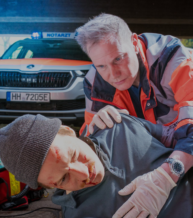 "Notruf Hafenkante - Wolle und Feines": Dr. Haase (Fabian Harloff) kniet hinter Jannis Frings (Merlin Sandmeyer), der verletzt auf dem Boden liegt. Seine linke Hand liegt auf der Brust des Verletzten, die rechte liegt auf dessen Schulter. Im Hintergrund steht ein Notarztwagen und daneben ein Rettungsassistent (Komparse). Neben Dr. Haase liegt sein geöffneter Erste-Hilfe-Koffer.