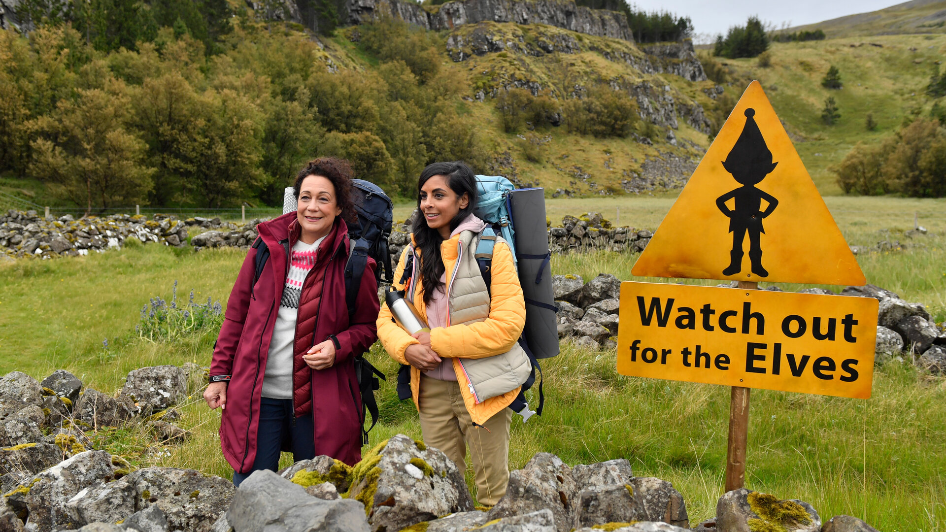 "Das Traumschiff: Island": In einer nordischen Naturkulisse: Auf einer grünen Wiese stehen Hanna Liebhold (Barbara Wussow) und Dr. Jessica Delgado (Collien Fernandes) nebeneinander und blicken lachend in die Ferne. Sie tragen dicke Jacken und Wanderrucksäcke. Dr. Delgado hält eine Thermoskanne in den Händen. Vor ihnen und auch im Hintergrund sind moosbewachsene Steine zu kleinen Mauern aufgetürmt. Neben ihnen steht ein gelbes Warnschild mit dem Abbild eines Elf und der Aufschrift "Watch out for the Elves".