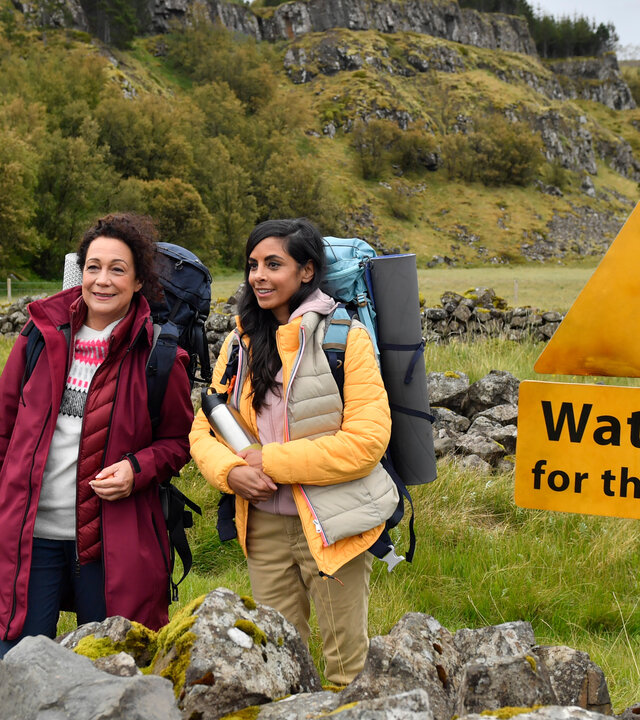 "Das Traumschiff: Island": In einer nordischen Naturkulisse: Auf einer grünen Wiese stehen Hanna Liebhold (Barbara Wussow) und Dr. Jessica Delgado (Collien Fernandes) nebeneinander und blicken lachend in die Ferne. Sie tragen dicke Jacken und Wanderrucksäcke. Dr. Delgado hält eine Thermoskanne in den Händen. Vor ihnen und auch im Hintergrund sind moosbewachsene Steine zu kleinen Mauern aufgetürmt. Neben ihnen steht ein gelbes Warnschild mit dem Abbild eines Elf und der Aufschrift "Watch out for the Elves".