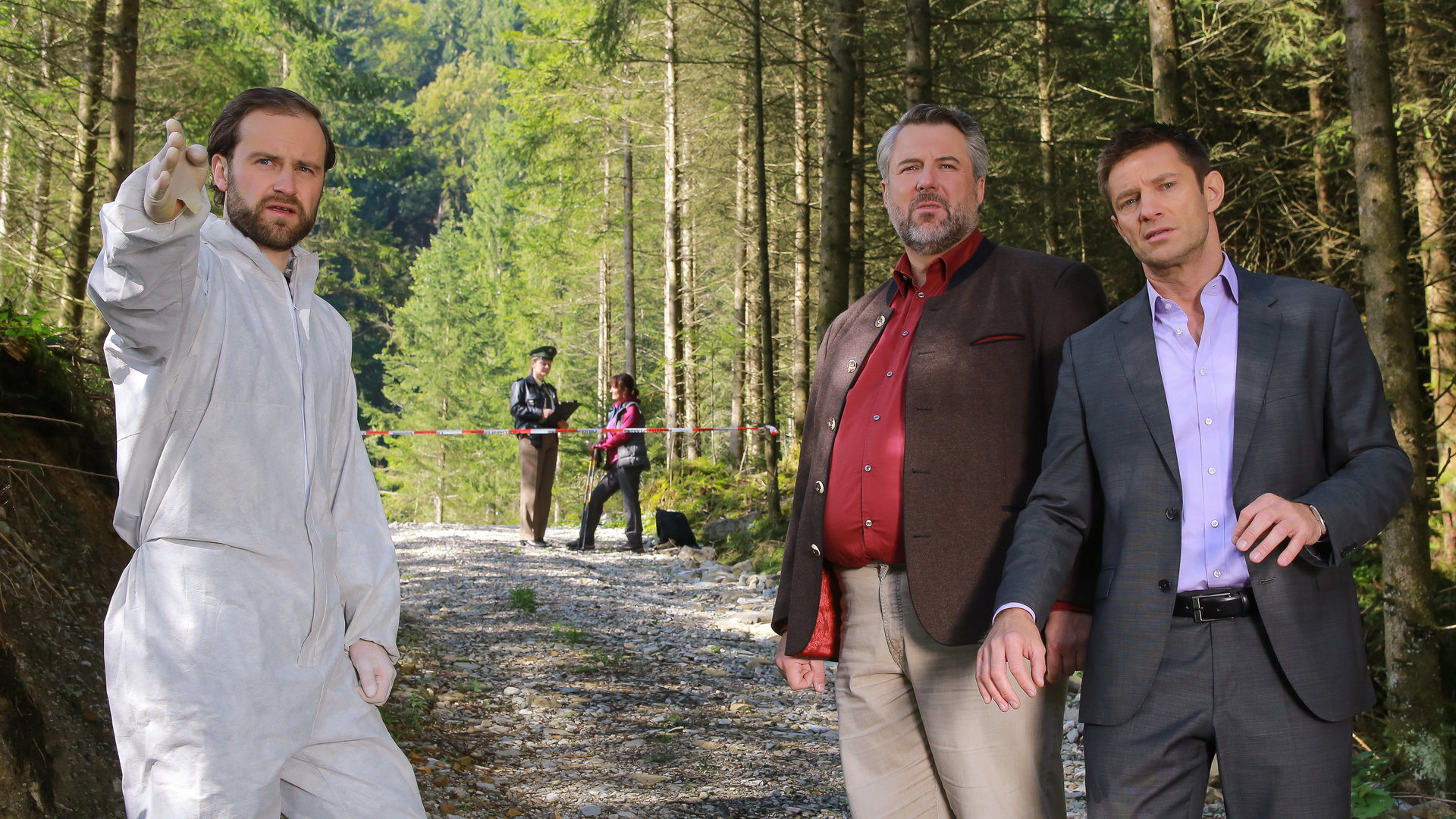 "Die Rosenheim-Cops - In die Falle gerast": Im Wald zeigt Spurensicherer Franz Leitner (Simon Sebastian Gerasch) in eine Richtung. Die Kommissare Stadler (Dieter Fischer) und Hansen (Igor Jeftic) stehen daneben und schauen in diese Richtung.