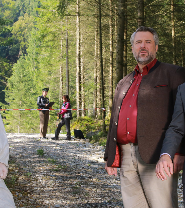 "Die Rosenheim-Cops - In die Falle gerast": Im Wald zeigt Spurensicherer Franz Leitner (Simon Sebastian Gerasch) in eine Richtung. Die Kommissare Stadler (Dieter Fischer) und Hansen (Igor Jeftic) stehen daneben und schauen in diese Richtung.