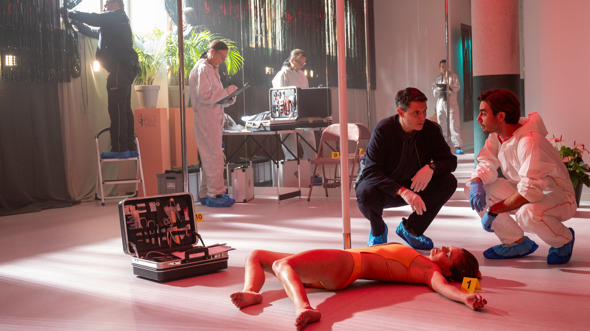 "SOKO Stuttgart - Poledance": Rico Sander (Benjamin Strecker) und Dr. Benedikt Förster (Florian Wünsche) hocken neben einer Poledance-Stange und blicken sich nachdenklich an. Beide tragen Gummihandschuhe und Fußüberzieher. Vor den Ermittlern liegt die Leiche von Lilly Martins (Komparsin). Im Hintergrund arbeiten drei Mitglieder der Spurensicherung (Komparsen) in weißen Schutzanzügen und ein Polizist (Komparse).
