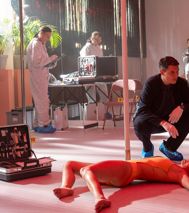 "SOKO Stuttgart - Poledance": Rico Sander (Benjamin Strecker) und Dr. Benedikt Förster (Florian Wünsche) hocken neben einer Poledance-Stange und blicken sich nachdenklich an. Beide tragen Gummihandschuhe und Fußüberzieher. Vor den Ermittlern liegt die Leiche von Lilly Martins (Komparsin). Im Hintergrund arbeiten drei Mitglieder der Spurensicherung (Komparsen) in weißen Schutzanzügen und ein Polizist (Komparse).