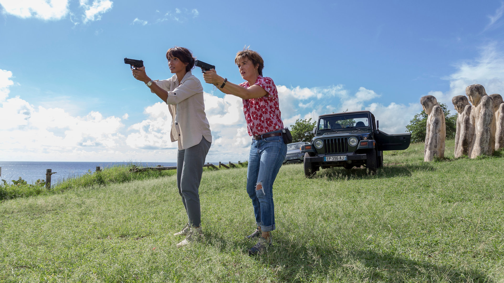 "Deadly Tropics - Späte Rache": Mélissa Sainte-Rose (Sonia Rolland) und Gaëlle Crivelli (Béatrice de la Boulaye) stehen nebeneinander auf einer Wiese am Meer und zielen mit ihren erhobenen Waffen in die Ferne.