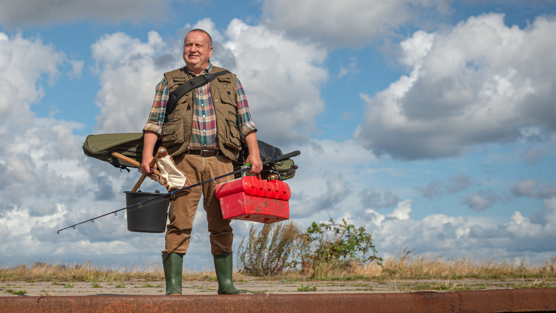 "SOKO Wismar – Breifrei statt Bierschinken": Reuter (Udo Kroschwald) steht in voller Angelmontur, mit Gummistiefeln und Angelweste, Eimer, Angel und Klappstuhl am Kai und blickt auf das vor ihm liegende Meer. Hinter ihm ist der leuchtendblaue Himmel mit unzähligen Wolken durchsetzt. Reuter hat einen zufriedenen Gesichtsausdruck.