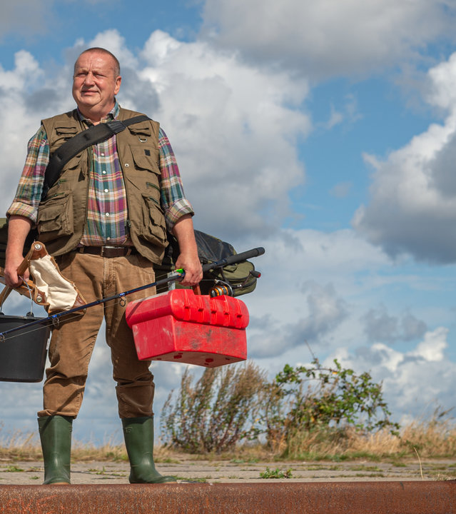 "SOKO Wismar – Breifrei statt Bierschinken": Reuter (Udo Kroschwald) steht in voller Angelmontur, mit Gummistiefeln und Angelweste, Eimer, Angel und Klappstuhl am Kai und blickt auf das vor ihm liegende Meer. Hinter ihm ist der leuchtendblaue Himmel mit unzähligen Wolken durchsetzt. Reuter hat einen zufriedenen Gesichtsausdruck.
