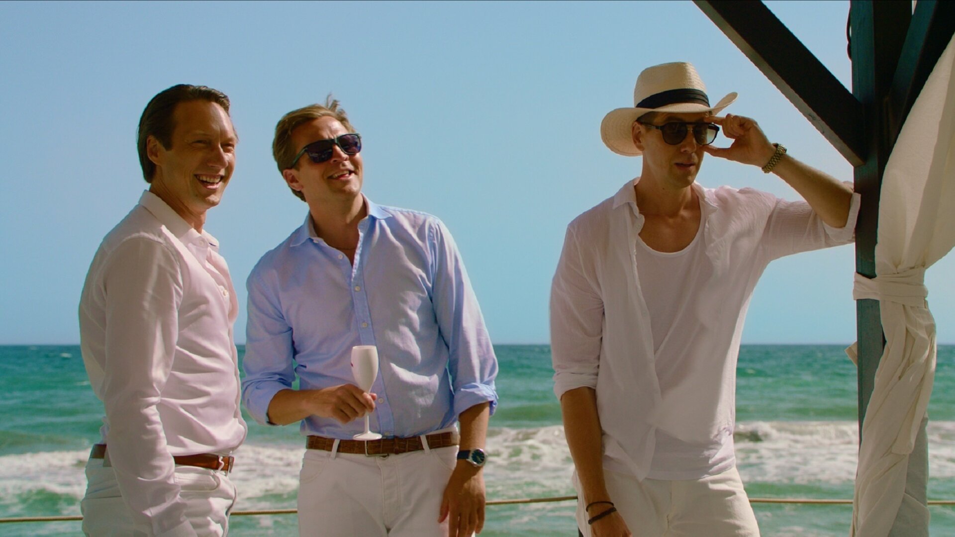 "Exit - Team play and Shadow play": Adam (Simon J. Berger), Henrik (Tobias Santelmann) und William (Pål Sverre Hagen) stehen gemeinsam lachend in der Sonne am Meer. Henrik hält ein Champagnerglas in der Hand. William lehnt sich an den Rahmen eines Strandbettes und greift dabei nach seiner Sonnenbrille.