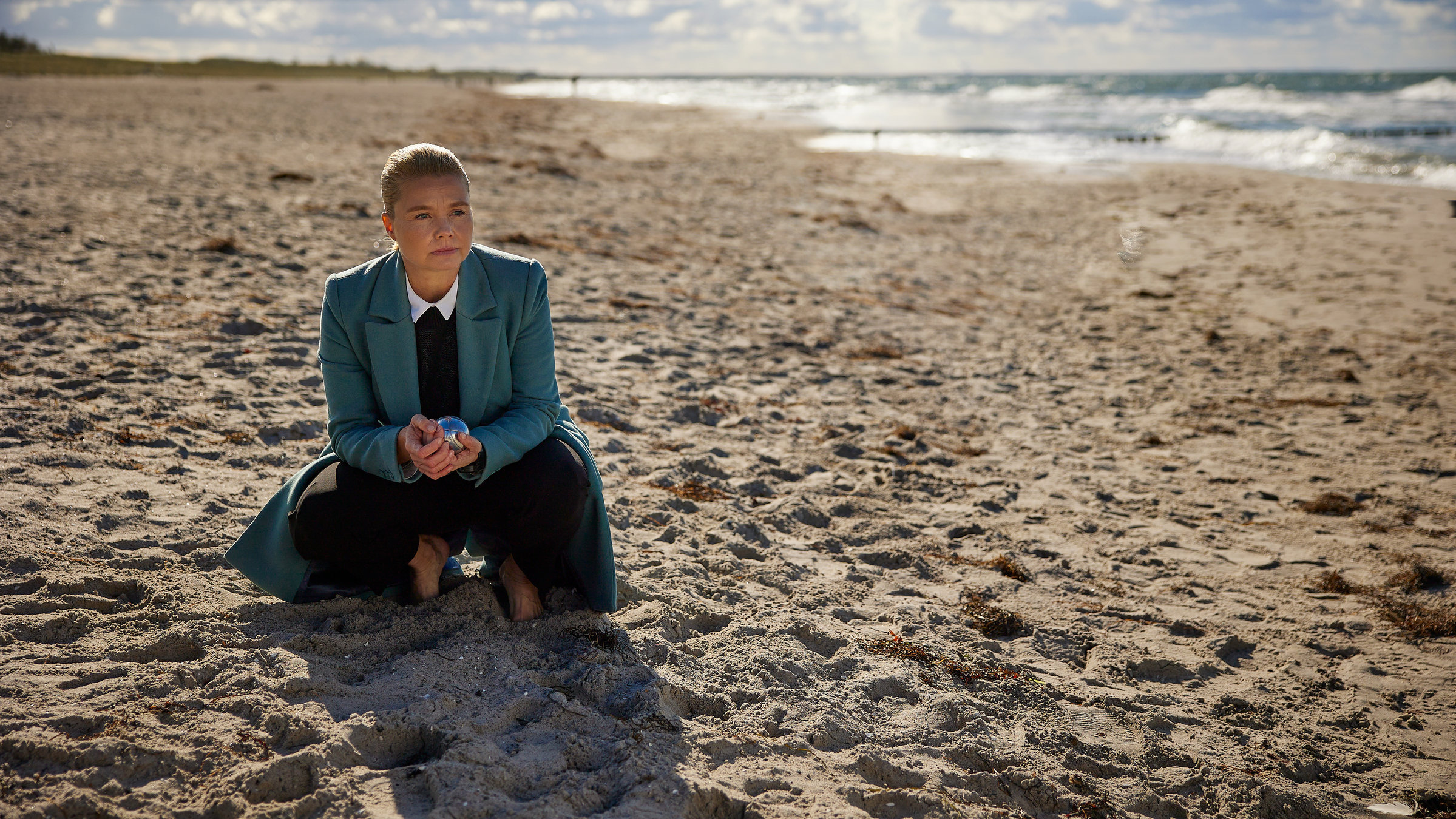 "Ella Schön - Das Glück der Erde": Ella (Annette Frier) kniet im Sand am Meer. In der Hand hält sie eine Boule-Kugel.
