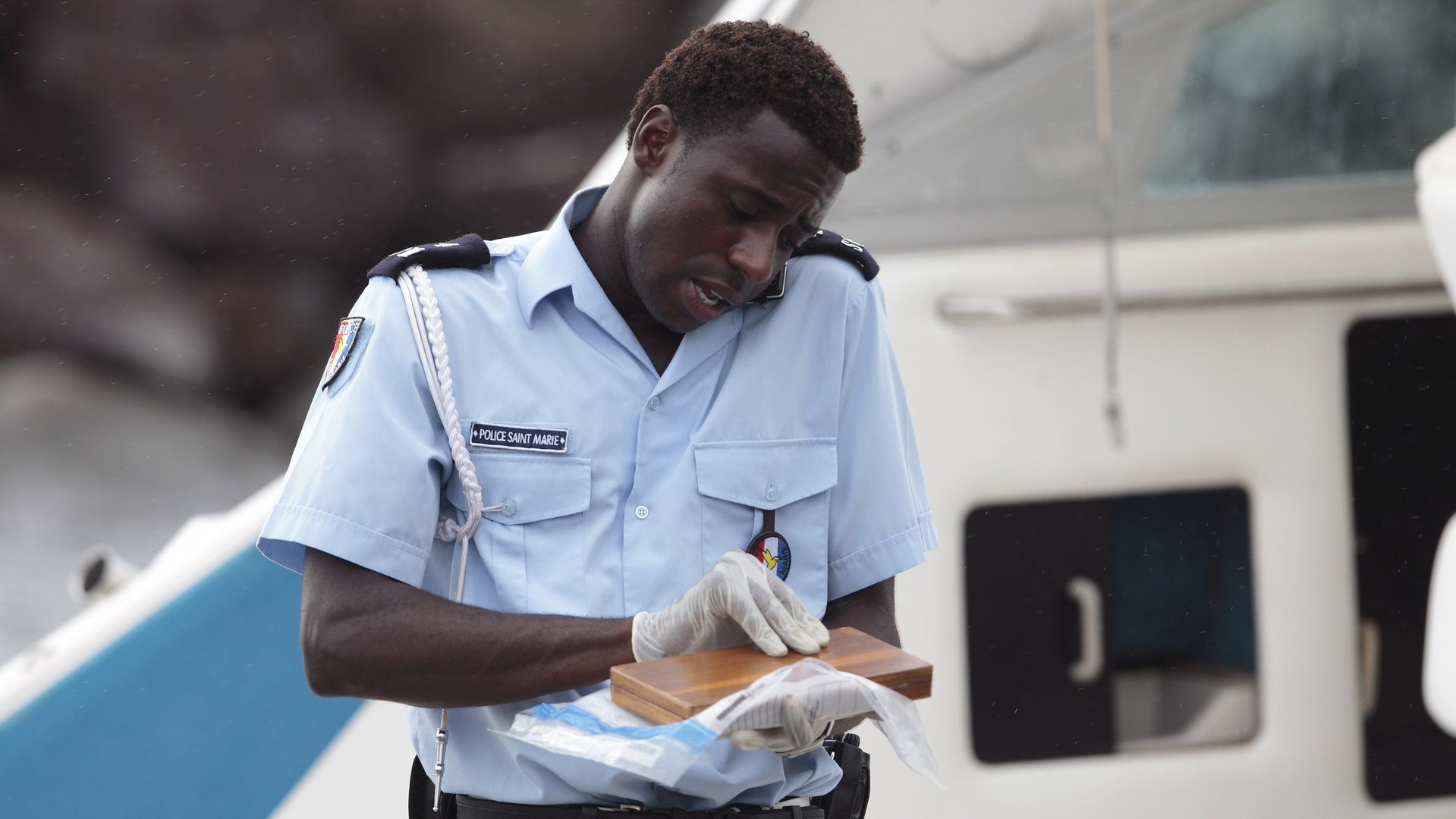 "Death in Paradise - Tropisches Fieber": Nahaufnahme Fidel (Gary Carr) auf einem Boot - er hält ein Handy zwischen Schulter und Kopf gepresst. In den Händen hält er eine kleine Holzschachtel.