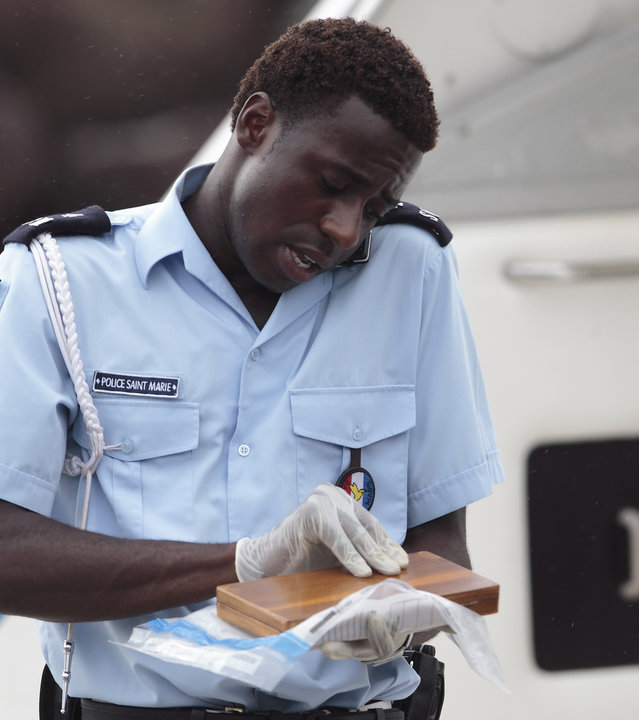 "Death in Paradise - Tropisches Fieber": Nahaufnahme Fidel (Gary Carr) auf einem Boot - er hält ein Handy zwischen Schulter und Kopf gepresst. In den Händen hält er eine kleine Holzschachtel.