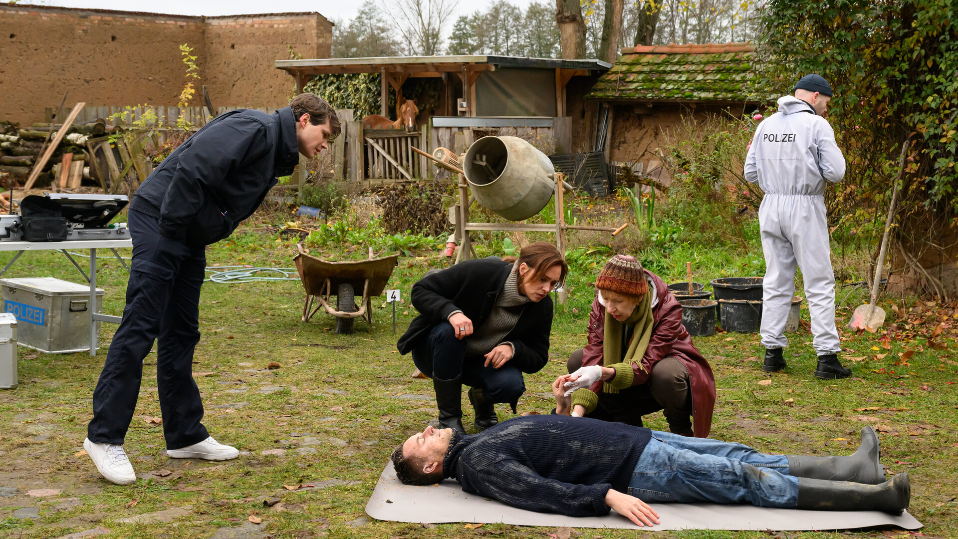 "SOKO Wismar - Unter Nachbarn": Im Hof auf einem Stück Wiese: Dr. Helene Sturbeck (Katharina Blaschke) zeigt Karoline Joost (Nike Fuhrmann) und Eddi Jansons (Gustavs Gailus) Wunden an der Leiche von Ben Kruse (Rico Höft).